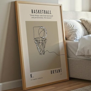 Könnte beinhalten: Ein gerahmter Basketball-Kunstdruck mit minimalistischem Design. Das Kunstwerk zeigt einen Basketball und einen Korb im Strichstil, mit dem Wort "BASKETBALL" oben. Der Druck befindet sich in einem hellfarbigen Rahmen, der an einer Wand in der Nähe eines Bettes lehnt.