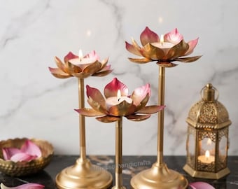 May include: Three gold-toned metal candle holders shaped like lotus flowers with pink-tipped petals, each holding a lit tea light candle. A small gold lantern with a lit candle and a bowl containing loose pink petals are also present.