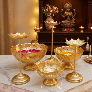 Puede incluir: Un conjunto de cuencos de metal dorado con forma de loto y portavelas elevados. Los cuencos presentan intrincados diseños de pétalos recortados y están llenos de pétalos de flores coloridos. Algunos soportes contienen velas encendidas.