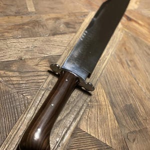 Op de afbeelding: Een groot Bowie-mes van gepolijst staal met een donkerhouten handvat. Het lemmet is lang en breed, met een scherpe punt. Het handvat is gebogen voor een comfortabele grip en is vastgezet met metalen stutten. Het mes ligt op een houten oppervlak.
