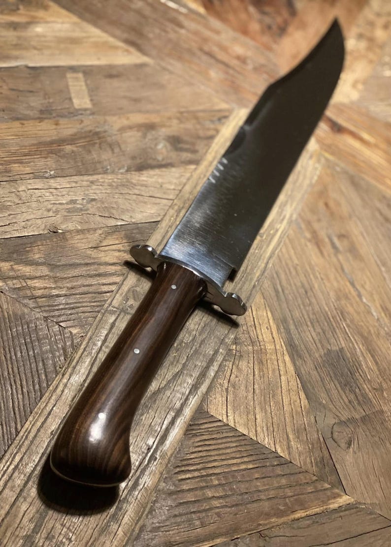 Op de afbeelding: Een groot Bowie-mes van gepolijst staal met een donkerhouten handvat. Het lemmet is lang en breed, met een scherpe punt. Het handvat is gebogen voor een comfortabele grip en is vastgezet met metalen stutten. Het mes ligt op een houten oppervlak.