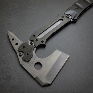 Pu&ograve; includere: Un'ascia tattica tomahawk con manico nero e lama grigia. L'ascia presenta un'impugnatura strutturata, una testa a martello e una lama affilata. Il design include un foro vicino alla lama e diverse viti per l'assemblaggio.
