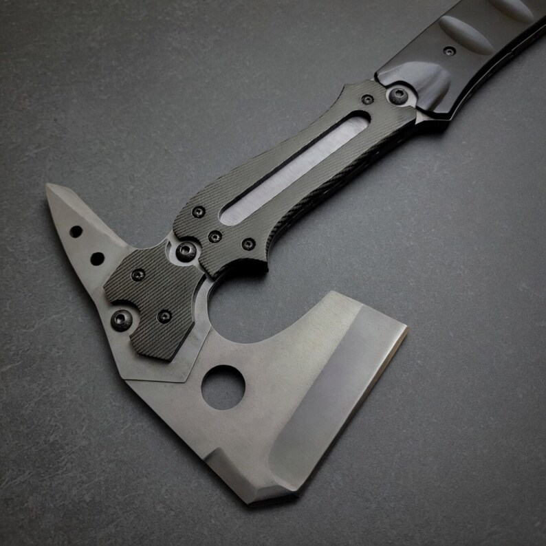 Pu&ograve; includere: Un'ascia tattica tomahawk con manico nero e lama grigia. L'ascia presenta un'impugnatura strutturata, una testa a martello e una lama affilata. Il design include un foro vicino alla lama e diverse viti per l'assemblaggio.