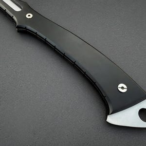 May include: Close-up of a black knife handle with a silver blade. The handle is curved and has a smooth, glossy finish. The blade has a hole near the end. The knife appears to be a folding knife.