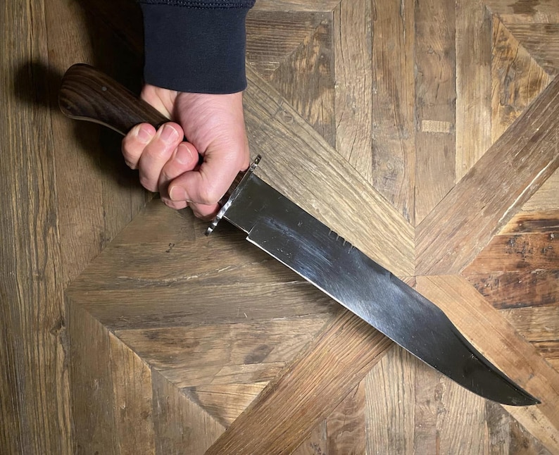 Op de afbeelding: Een groot roestvrijstalen mes met een donkerhouten handvat. Het lemmet is breed en heeft een licht gebogen rand. Het mes wordt in een hand gehouden, met het handvat naar de kijker gericht.