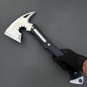 May include: A silver and black tactical axe with a sharp blade and a gear-shaped cutout. The axe features a pointed spike on the top and is held by a gloved hand against a dark gray background.
