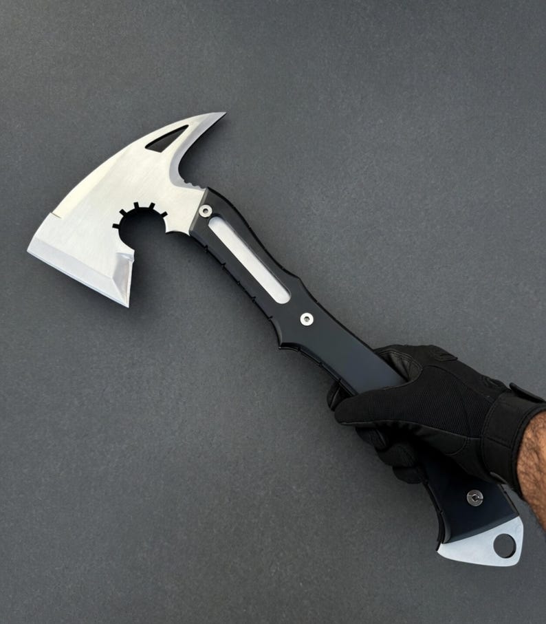 May include: A silver and black tactical axe with a sharp blade and a gear-shaped cutout. The axe features a pointed spike on the top and is held by a gloved hand against a dark gray background.