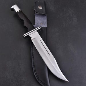 May include: A large, stainless steel knife with a curved blade and a black handle. The knife is partially inserted into a black leather sheath with a silver button closure. The handle has a silver bolster.