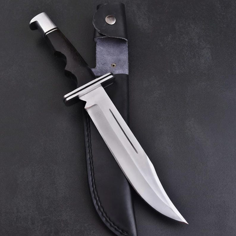 May include: A large, stainless steel knife with a curved blade and a black handle. The knife is partially inserted into a black leather sheath with a silver button closure. The handle has a silver bolster.