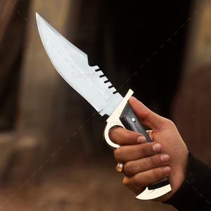 May include: A large, silver-bladed knife with a serrated edge and a black wooden handle. The knife features a gold-colored finger ring and accents. The blade is sharp and the handle is textured for grip.
