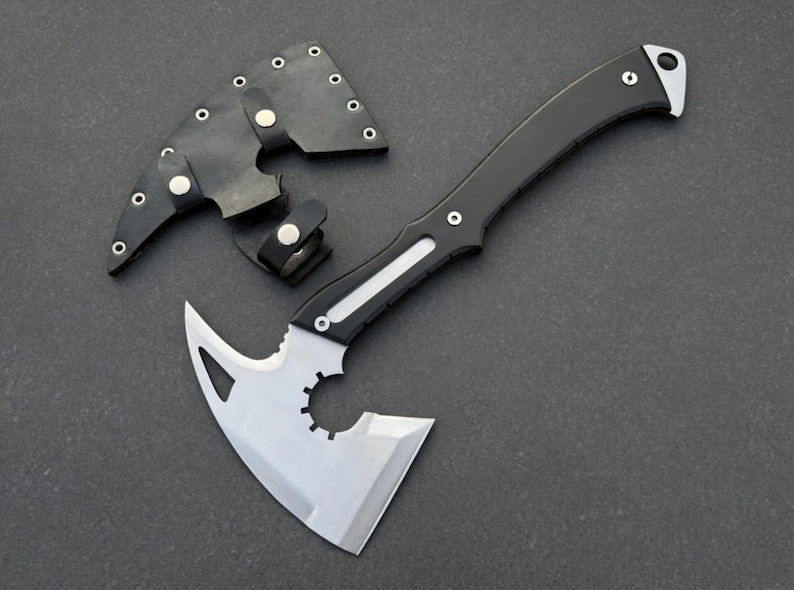 May include: A black and silver hatchet with a curved handle and a protective leather sheath. The hatchet blade has a unique design with a cutout and a sharp edge. The sheath is black with silver rivets.