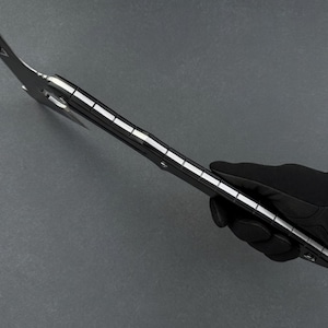 May include: A black and silver multi-tool axe held by a gloved hand. The axe head has a curved blade and a pointed end. The handle is black with silver accents. The tool is designed for various outdoor and survival tasks.