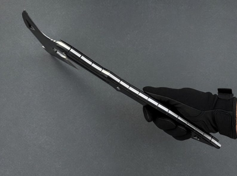 May include: A black and silver multi-tool axe held by a gloved hand. The axe head has a curved blade and a pointed end. The handle is black with silver accents. The tool is designed for various outdoor and survival tasks.