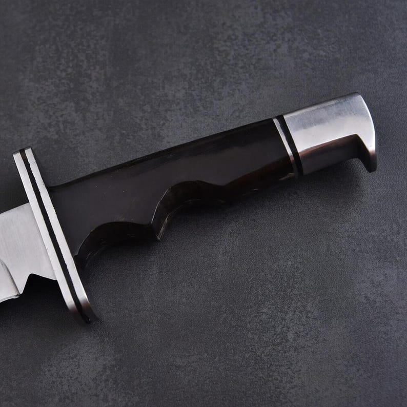 May include: Close-up of a hunting knife with a black handle and a silver blade. The handle has a curved design for a secure grip. The blade is partially visible, showing a sharp edge and a silver guard.