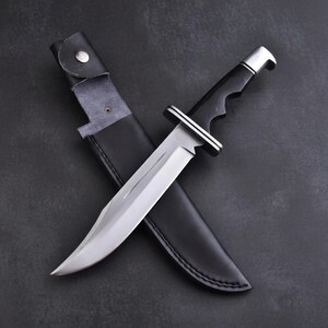 May include: A large, stainless steel knife with a curved blade and a black handle. The knife is partially sheathed in a black leather case. The handle has a silver bolster. The knife is designed for outdoor use.