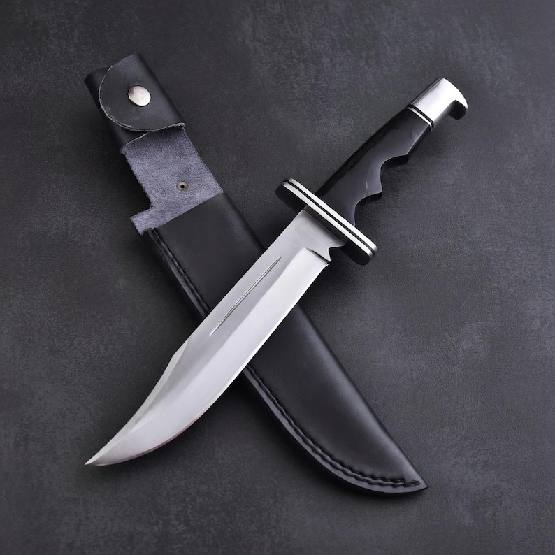 May include: A large, stainless steel knife with a curved blade and a black handle. The knife is partially sheathed in a black leather case. The handle has a silver bolster. The knife is designed for outdoor use.