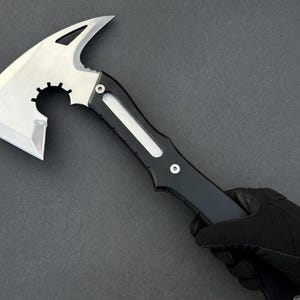 May include: A tactical tomahawk axe with a silver blade and a black handle. The axe head features a curved blade, a pointed spike, and a circular cutout. The handle has a streamlined design with a white accent.
