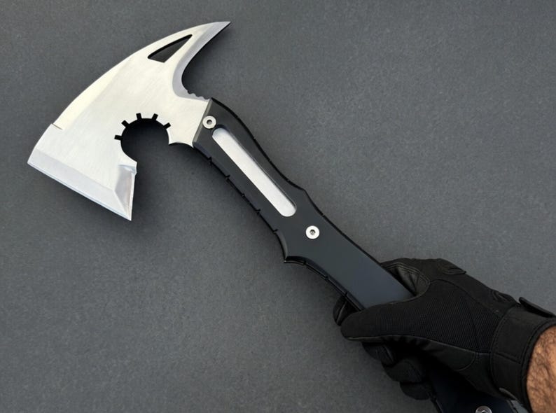 May include: A tactical tomahawk axe with a silver blade and a black handle. The axe head features a curved blade, a pointed spike, and a circular cutout. The handle has a streamlined design with a white accent.