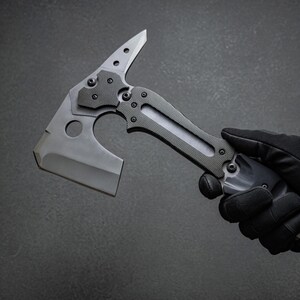 Pu&ograve; includere: Un tomahawk tattico con finitura grigia. La testa dell'ascia presenta una lama affilata, una punta e un ritaglio circolare. L'impugnatura &egrave; strutturata per l'aderenza e ha una combinazione di colori grigio e nero. L'ascia &egrave; tenuta da una mano guantata.
