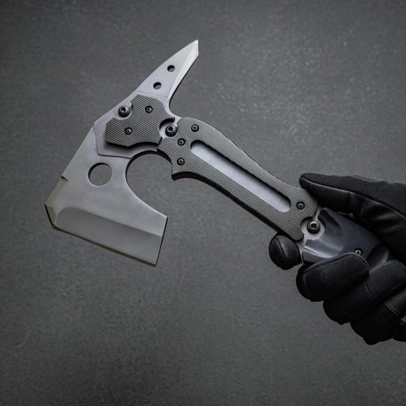 Pu&ograve; includere: Un tomahawk tattico con finitura grigia. La testa dell'ascia presenta una lama affilata, una punta e un ritaglio circolare. L'impugnatura &egrave; strutturata per l'aderenza e ha una combinazione di colori grigio e nero. L'ascia &egrave; tenuta da una mano guantata.