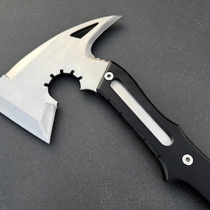 May include: A tactical axe with a silver blade and a black handle. The axe head features a curved blade, a pointed spike, and a circular cutout. The handle has a streamlined design with a white accent.