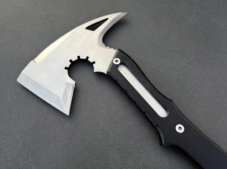 May include: A tactical axe with a silver blade and a black handle. The axe head features a curved blade, a pointed spike, and a circular cutout. The handle has a streamlined design with a white accent.