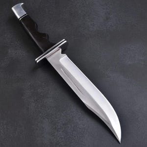 May include: A stainless steel hunting knife with a black handle and a silver bolster. The blade is curved and has a sharp edge. The knife is set against a dark background.