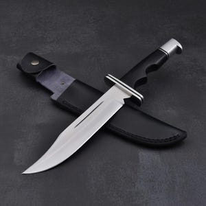 May include: A large hunting knife with a polished steel blade and a black handle. The knife is resting in a black leather sheath. The handle has a silver bolster and a finger groove for a secure grip.