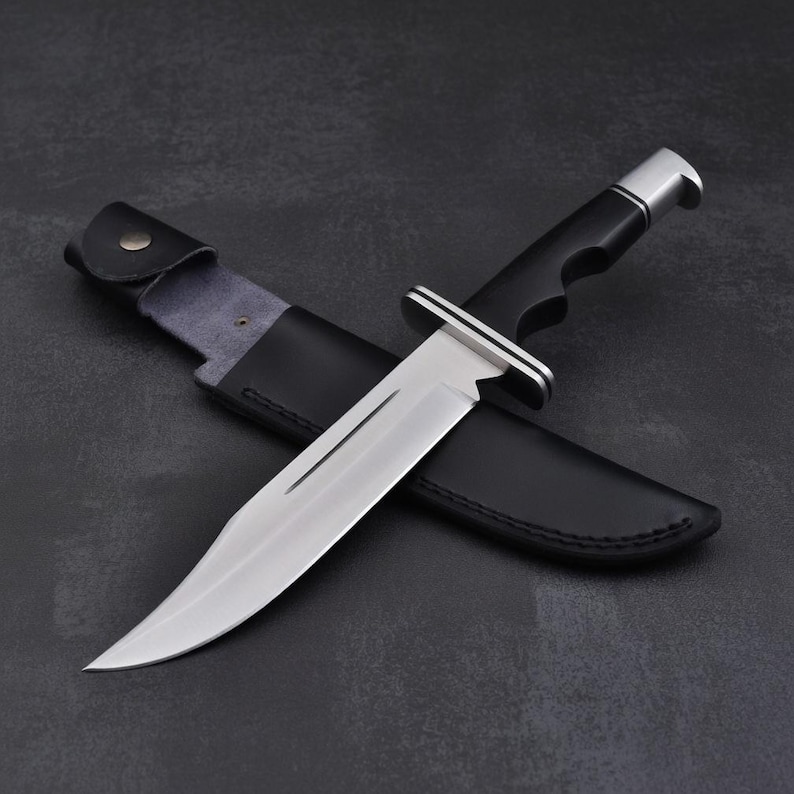 May include: A large hunting knife with a polished steel blade and a black handle. The knife is resting in a black leather sheath. The handle has a silver bolster and a finger groove for a secure grip.