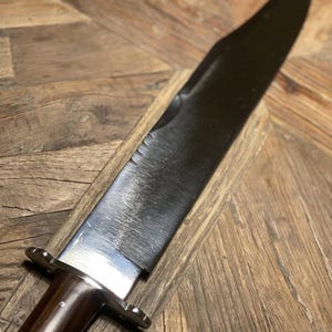 Op de afbeelding: Een groot, gepolijst mes met een donker lemmet en een houten handvat. Het lemmet heeft een gekartelde sectie bij het handvat. Het handvat is donkerbruin en heeft een metalen bolster. Het mes wordt getoond op een houten oppervlak.