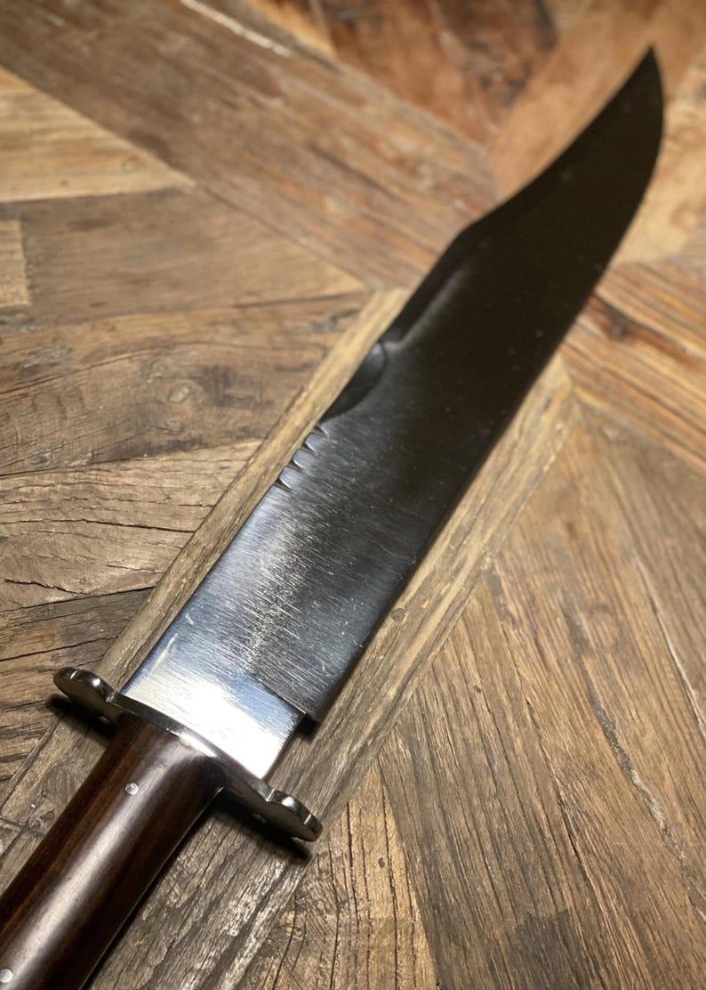 Op de afbeelding: Een groot, gepolijst mes met een donker lemmet en een houten handvat. Het lemmet heeft een gekartelde sectie bij het handvat. Het handvat is donkerbruin en heeft een metalen bolster. Het mes wordt getoond op een houten oppervlak.