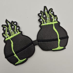 Pode incluir: Patch bordado preto com um design de sutiã estilizado com duas taças arredondadas conectadas por uma faixa. Cada taça tem um contorno verde e um design verde fluindo com detalhes brancos, assemelhando-se a uma poção borbulhante ou planta.