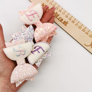 May include: A collection of handmade hair bows in various pastel colors, including pink, white, and blue. Each bow features a glittery finish and embroidered initials. A wooden ruler with the text "Wlif.co.uk" is in the background.