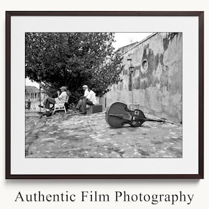 May include: Black and white framed photograph featuring a double bass resting on a cobblestone street. Two people are seated nearby, with a third person sitting on a bench. The photograph is labeled "Authentic Film Photography".
