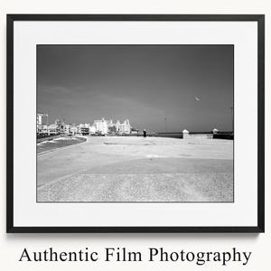 May include: Black and white framed photograph of a coastal scene. The image features a person walking on a paved area, with buildings in the background. The sky is clear, and the photograph is labeled "Authentic Film Photography."