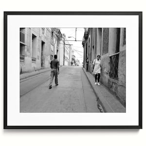 May include: A black and white framed photograph depicting a narrow urban street scene. A person walks away from the camera in the center of the road, while another person walks toward the camera along the sidewalk beside weathered stone buildings.