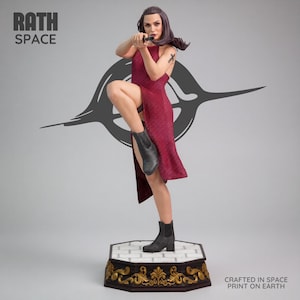May include: A figurine of a woman in a red dress and black boots, holding a gun. The figurine is standing on a decorative base with the text "RATH SPACE" and "CRAFTED IN SPACE PRINT ON EARTH".
