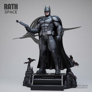 May include: A gray Batman statue holding a weapon, with a black cape and cowl. The statue stands on a black platform with bat-shaped accents. The text "RATH SPACE" is visible in the upper left corner.