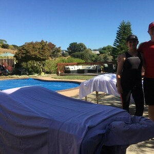 Puede incluir: Dos personas est&aacute;n de pie junto a una piscina con dos mesas de masaje cubiertas con s&aacute;banas blancas. La persona de la izquierda lleva un top negro sin mangas y pantalones negros. La persona de la derecha lleva una camiseta roja y pantalones cortos negros. La piscina es azul y hay una terraza de madera en el fondo.