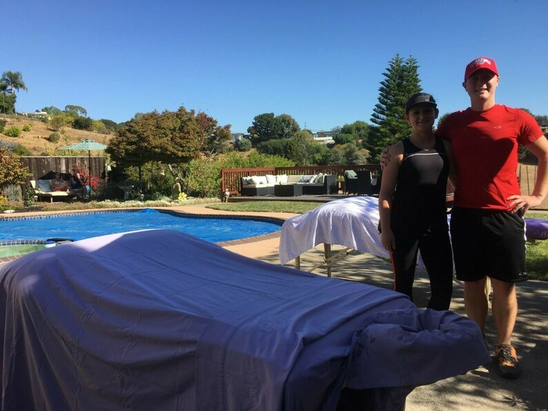 Puede incluir: Dos personas est&aacute;n de pie junto a una piscina con dos mesas de masaje cubiertas con s&aacute;banas blancas. La persona de la izquierda lleva un top negro sin mangas y pantalones negros. La persona de la derecha lleva una camiseta roja y pantalones cortos negros. La piscina es azul y hay una terraza de madera en el fondo.