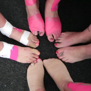 Puede incluir: Cinco pies con cinta atl&eacute;tica rosa y blanca envuelta alrededor de los tobillos. La cinta est&aacute; dise&ntilde;ada para sostener los tobillos durante la actividad f&iacute;sica.