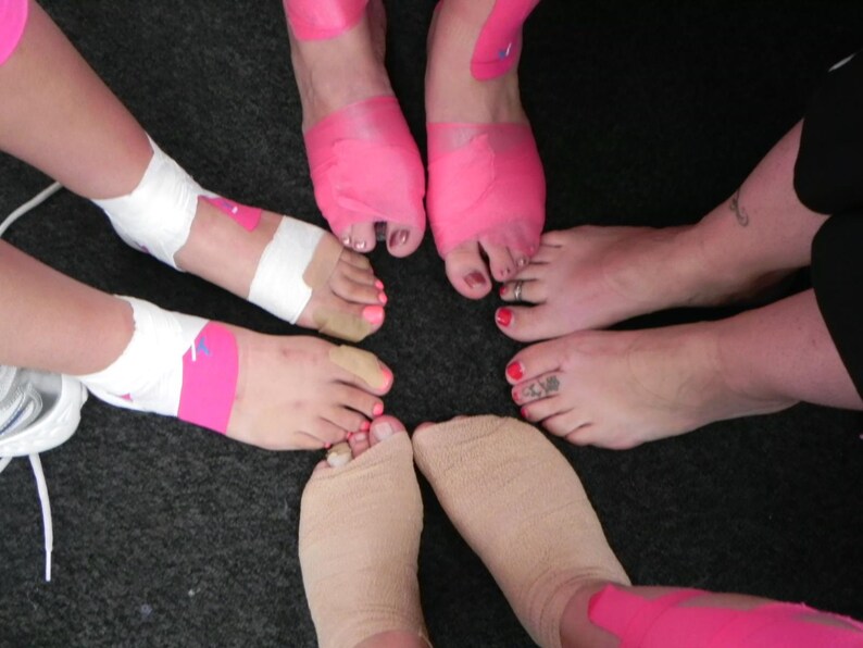 Puede incluir: Cinco pies con cinta atl&eacute;tica rosa y blanca envuelta alrededor de los tobillos. La cinta est&aacute; dise&ntilde;ada para sostener los tobillos durante la actividad f&iacute;sica.