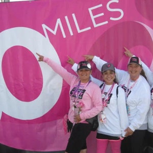 以下が含まれることがあります： ピンクのシャツと帽子をかぶった4人の女性が、白文字で「30 MILES」と書かれたピンクのバナーの前に立っています。女性たちは笑顔で、レースやチャリティイベントに参加しているようです。