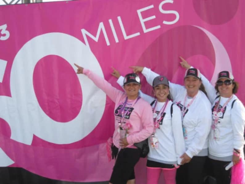 以下が含まれることがあります： ピンクのシャツと帽子をかぶった4人の女性が、白文字で「30 MILES」と書かれたピンクのバナーの前に立っています。女性たちは笑顔で、レースやチャリティイベントに参加しているようです。