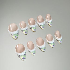 May include: Set of ten artificial nails. Each nail features a light beige oval tip, a white fluffy trim, and a floral design with purple flowers on a yellow background. The nails are arranged on a reflective surface.
