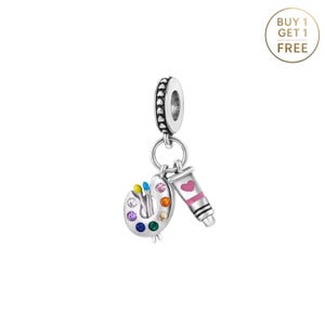 May include: A silver charm featuring a painter's palette with colorful paint and brushes, and a tube of paint with a pink heart. The charm has a decorative top with a ring for attachment. The text "BUY 1 GET 1 FREE" is in a circle.