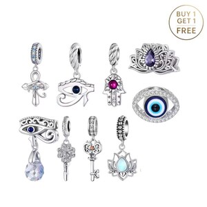 May include: A collection of silver-toned charm pendants with various designs, including an ankh, an eye of Horus, a Hamsa hand, keys, and a lotus flower. Some charms feature colored gemstones and crystals. The image includes a "Buy 1 Get 1 Free" circular graphic.