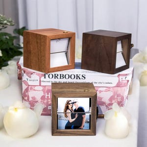May include: Three wooden photo display boxes in varying shades of brown. One box displays a photo of a couple. The boxes are square with a rectangular opening. The boxes are sitting on top of a book with the title "TORBOOKS".