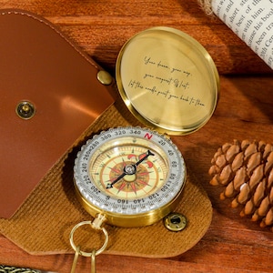 Peut inclure: Une boussole en laiton vintage avec un étui en cuir. La boussole a un cadran détaillé avec des indications directionnelles et une aiguille. Le couvercle de la boussole porte une citation gravée. L'étui est marron et la boussole est sur une surface en bois.