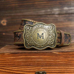 Op de afbeelding: Een bruine leren riem met een decoratieve messing gesp. De gesp heeft een gegraveerd ontwerp met het woord "Moroco" en de letter "M". De riem heeft een bloemenpatroon en ligt op een houten kist.
